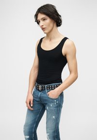 Νεαρός με μεσαίου μήκους σκούρα μαλλιά, που φοράει μαύρο αμάνικο τοπ, σκισμένο μπλε τζιν και μαύρη ζώνη με ασημί τρύπες.