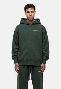 Felpa verde con zip realizzata in tessuto morbido, dotata di tasche a kangaroo. Presenta una stampa del logo frontale e un cappuccio con coulisse. Vestibilità classica.