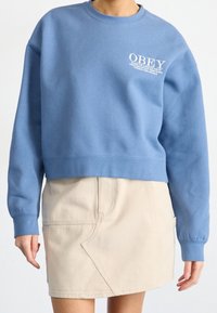 Blå bomullströja med rundad halsringning och vit "OBEY"-logotyp på bröstet, i kombination med en beige kjol med vinklade sömmar.