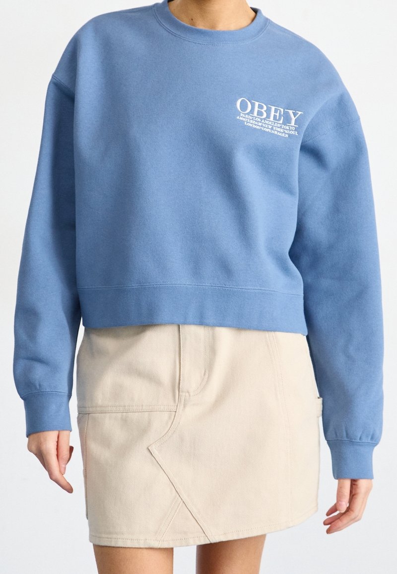 Blå bomullströja med rundad halsringning och vit "OBEY"-logotyp på bröstet, i kombination med en beige kjol med vinklade sömmar.