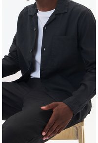 Czarna koszula z guzikami, o luźnym kroju, z naszywaną kieszenią i klasycznym kołnierzykiem, noszona na białym podkoszulku; pokazana z czarnymi spodniami.