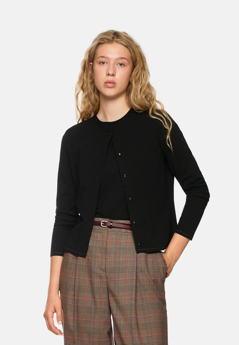 Schwarzer Cardigan mit Rundhalsausschnitt, vorderer Knopfverschluss und langen Ärmeln. Getragen über einem schwarzen Oberteil und karierter, hochtaillierter Hose.