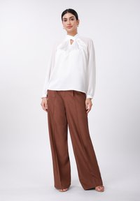 Blouse blanche avec un détail à découpe, manches longues, associée à un pantalon large marron. Tissu lisse, coupe ample et léger éclat subtil.