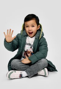 Groene pufferjack met capuchon, gelaagd over een witte graphic T-shirt. Grijze jeans en witte sneakers met groene accenten. Zittend en zwaaiend.