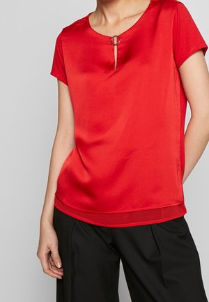 Kobieta ubrana w czerwoną bluzkę z krótkim rękawem z okrągłą klamrą przy dekolcie oraz czarne spodnie, stojąca z jedną ręką w kieszeni.