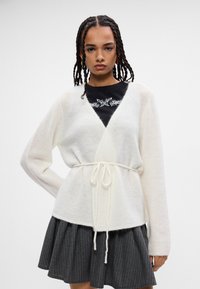 Měkký bílý cardigan s texturovaným povrchem a zavazováním v pase. Dlouhé rukávy, v kombinaci s tmavou plisovanou sukní s proužkovaným vzorem.