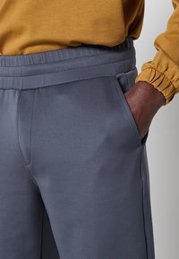 Pantaloni sportivi grigi con una texture liscia, vita elastica e tasche laterali, abbinati a una maglietta a maniche lunghe color senape.