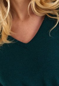 Pull vert à col en V avec une texture douce, présentant un motif tricot subtil et une coupe confortable. L'encolure est finie avec un bord côtelé.