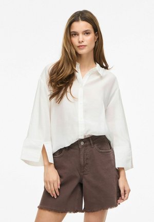 Žena s dlouhými hnědými vlasy má na sobě volnou bílou košili na knoflíky a hnědé džíny s třepeněným lemem, stojí před bílým pozadím.