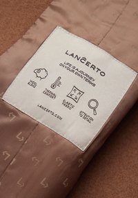 Etykieta z tkaniny mieszanki wełnianej w delikatnym brązie z ikonami oznaczającymi komfort termiczny, elastyczny materiał i dbałość o szczegóły, z wytłoczonym logo.