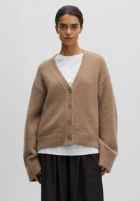 Beige wollen cardigan met een v-hals en vijf knopen. Heeft een losse pasvorm, geribde zoom en oversized mouwen. Gedragen over een wit shirt.