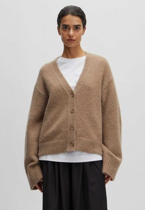 Beige wollen cardigan met een v-hals en vijf knopen. Heeft een losse pasvorm, geribde zoom en oversized mouwen. Gedragen over een wit shirt.