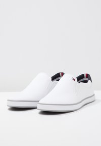 Scarpe slip-on bianche con tomaia in canvas liscia, suola in gomma grigia e interno navy a contrasto con un piccolo dettaglio logo rosso e bianco.