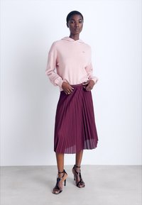 Sudadera rosa claro con cordón, combinada con una falda midi plisada burdeos. La modelo usa tacones negros con tiras, de pie sobre un fondo neutro.