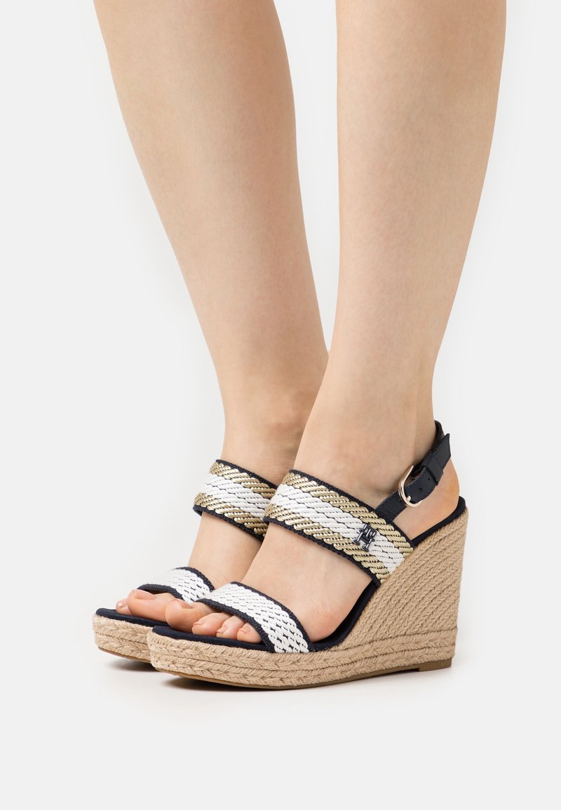 Pieds portant des sandales compensées tissées beige et blanches avec brides à la cheville et fermetures à boucle sur un fond neutre.
