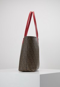 Brun mönstrad tygväska med röda läderremmar, med en strukturerad design och texturerad yta. Inga synliga h årdvaror eller detaljer.