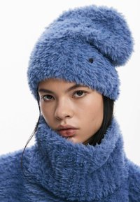 Udune sinine müts ümarate toppidega ja tupsuga, valmistatud pehmest materjalist; kaunistatud musta nupuga. Sobiv udune kaelus.