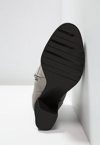 Semelle en caoutchouc noir d'une bottine grise à talon bloc reposant contre un mur blanc sur un sol blanc.