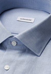 Close-up van een lichtblauwe overhemdenkraag met witte knopen en een wit 'seidensticker' merklabel binnenin de halslijn.