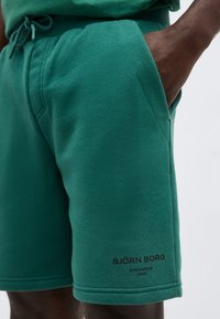 Άτομο με σκουρόχρωμο δέρμα που φοράει πράσινα σορτς Björn Borg, με το χέρι στην τσέπη, ορατό λογότυπο και ελαστική μέση με κορδόνι.