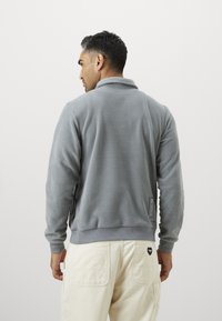 Γκρι φλις μπουφάν με ψηλό γιακά, πλεκτές μανσέτες και ζώνη. Διαθέτει μικρή λεπτομέρεια λογότυπου στο πλάι και απαλή υφή.