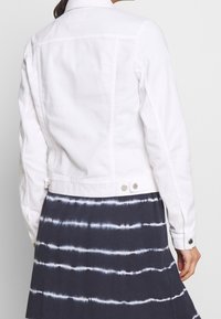 Vit denimjacka med en figursydd design, bakre söm, knäppning med knappar och justerbara sidor. Bärs över en blårandig tie-dye kjol.