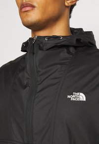 Close-up van een persoon in een zwart gewatteerd jack met een rits op de borst en het witte The North Face-logo aan de linkerkant.