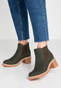 Botas de tobillo en verde oliva, hechas de ante, con paneles laterales elásticos, un tacón grueso en color marrón claro y una suela de goma texturizada para mayor agarre.