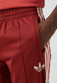 Röda träningsbyxor med resårmidja, krämfärgade sidostraper och en vit Adidas-logotyp på underbenet. Slät tygtextur.