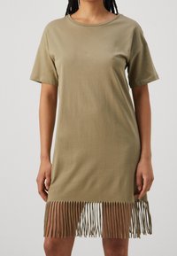 Kvinna i en kortärmad olivgrön klänning med fransdetaljer längs fållen mot en enhetlig bakgrund.