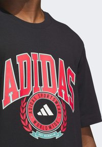 Černé bavlněné tričko s velkým červeným logem "ADIDAS", kruhovou nášivkou s bílým trojlístkem a vavřínovými akcenty. Standardní provedení s kulatým výstřihem.