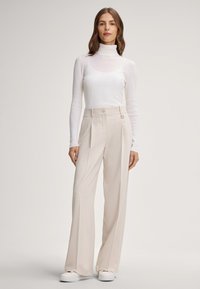 Weißer gerippter Rollkragenpullover kombiniert mit beigen weit geschnittenen Hosen mit Knopfdetail, getragen mit weißen Sneakers. Einfaches und modernes Design.