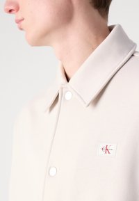 Giacca color beige chiaro con colletto strutturato, bottoni a scatto e una patch con logo che presenta scritte rosse su uno sfondo bianco quadrato.