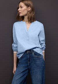Blouse bleu clair à manches longues avec un col en V et des poignets retroussés, associée à un jean denim bleu foncé doté de poches avant et d'une coupe classique.