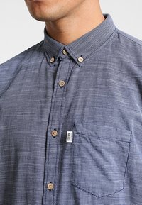 Knappskjorta i marinblå och grå med rutigt mönster. Har en krage, träknappar och en vänster bröstficka med en etikett.