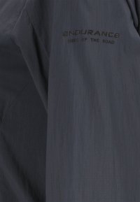 Marinblå lättviktig jacka med en subtil textur, med "ENDURANCE" och "HERO OF THE ROAD" tryckt i svart på ärmen.