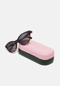 Óculos de sol estilo gato de tartaruga com lentes escuras ao lado de uma capa dura em dois tons, rosa e verde. A capa apresenta detalhes em dourado e o nome da marca.