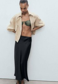 Veste beige cropped, top bandeau vert, jupe maxi en satin noir, délicate chaîne en or avec des détails floraux, textures lisses, design contemporain.