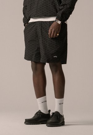 Pantalones cortos negros con estampados, fabricados en tejido liviano, combinados con calcetines blancos y zapatos negros. Destacan los detalles del logo y el corte relajado.