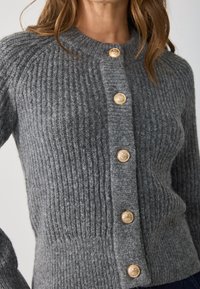 Gray knitted cardigan featuring a ribbed texture, round neck, and six goldtone buttons down the front. Soft material and fitted shape.