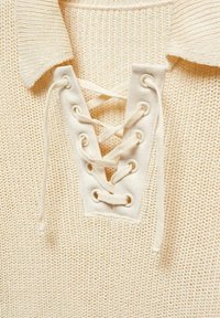 Lace-up knit top in cream color with a textured fabric, featuring a V-neck design, white cord laces, and circular metal eyelets.