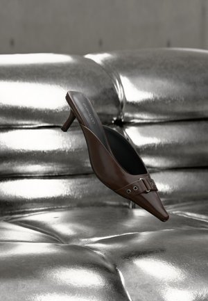 Mule à talon brun à bout pointu avec détail boucle flottant au-dessus d'une surface en cuir argenté capitonnée brillante.