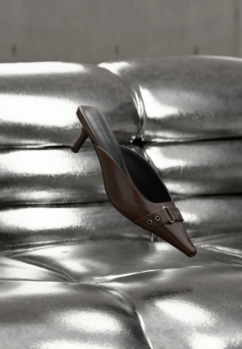 Καφέ μυτερή λουρίδα mule με τακούνι και λεπτομέρεια αγκράφας που αιωρείται πάνω από μία λαμπερή ασημί δερμάτινη επιφάνεια με μπαλώματα.