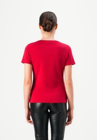 Woman with hair in a bun wearing a red short-sleeve shirt and black leather pants, standing facing away against a plain white background.