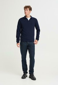 Jonge man met krullend haar, gekleed in een marineblauwe trui, een wit onderhemd, donkere jeans en zwarte schoenen, staande tegen een lichte achtergrond.