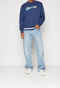 Marinblå sweatshirt med vit och aqua "AIR"-logotyp, kombinerad med ljusblå jeans och vita sneakers. Enkel design med rund halsringning.