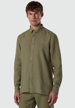 Giovane con capelli ricci che indossa una camicia verde oliva a maniche lunghe con bottoni e pantaloni abbinati, in piedi davanti a uno sfondo neutro.