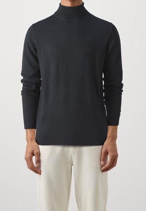 Homme portant un pull noir à col roulé et un pantalon clair, debout, les bras détendus le long du corps.