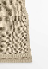 Gilet en maille beige sans manches avec motif en maille ouverte et ourlet côtelé sur fond blanc, montrant la moitié inférieure du vêtement.