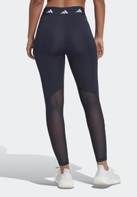 Leggings da allenamento di colore blu navy con pannelli in rete sui lati, una cintura ampia con tre strisce e scarpe da ginnastica bianche.